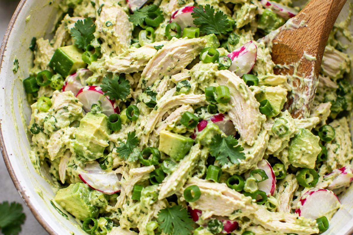 Creamy Avocado Chicken Salad Recipe with Lime and Cilantro 3 Close overhead view of avocado chicken salad recipe being gently folded in a bowl, showing chicken, red radish pieces, green scallions, cilantro, and creamy avocado dressing
