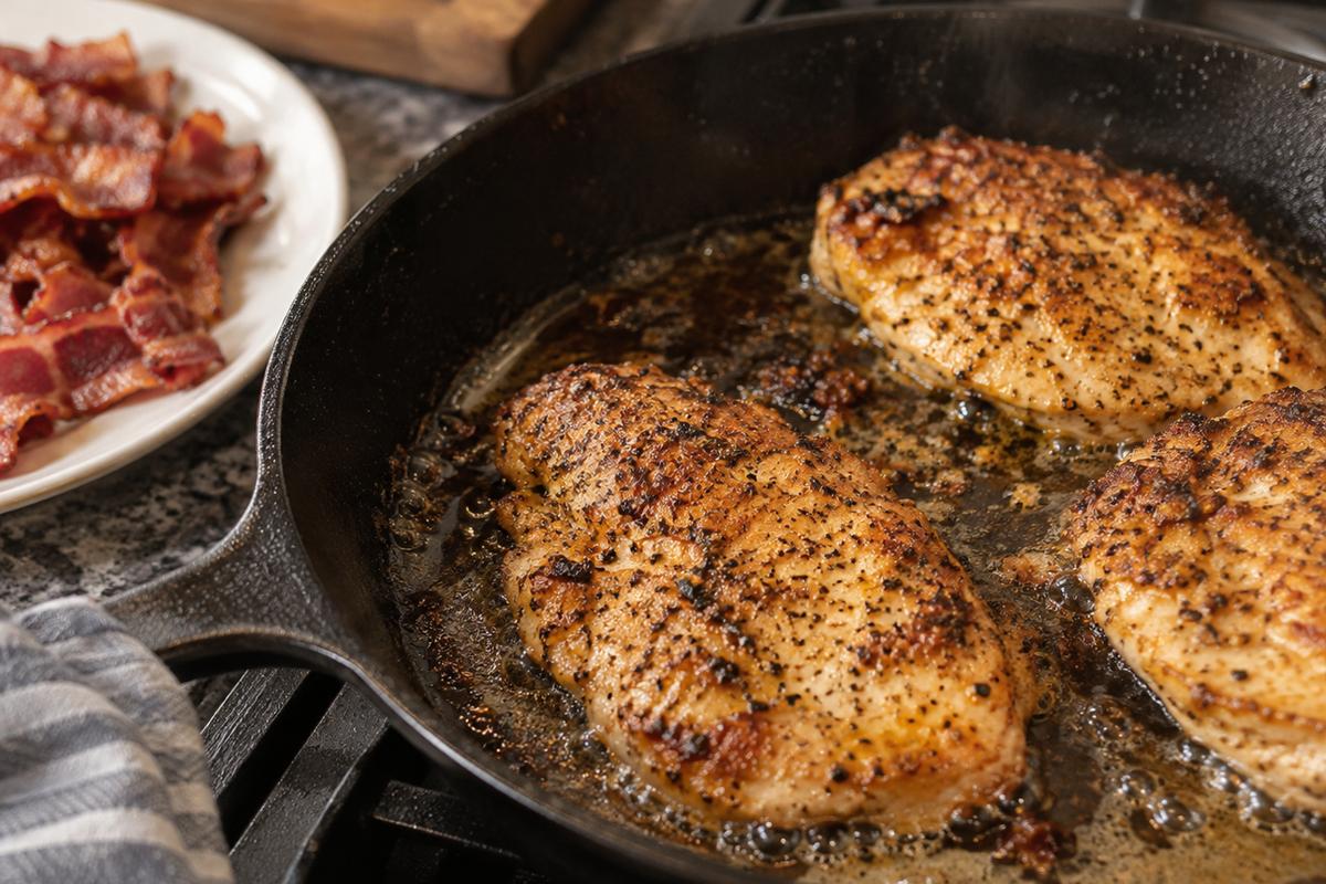 A close skillet view of creamy chicken bacon ranch pasta being prepared, showing golden seasoned chicken cutlets searing in a black skillet with a small amount of bacon drippings and crisp bacon pieces waiting on a plate beside the pan