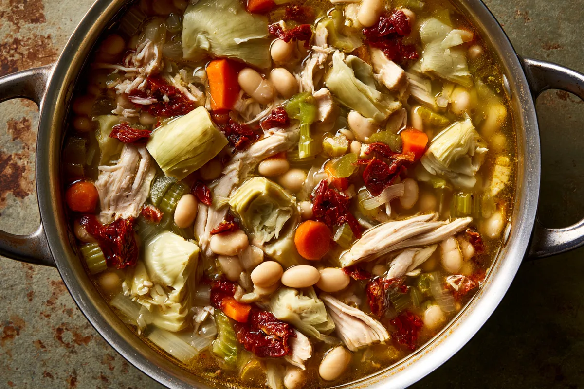High Fiber Chicken Soup with White Beans and Artichokes 3 A simmering pot of high fiber chicken soup with shredded chicken, white beans, chopped artichoke hearts, carrots, celery, and sun-dried tomatoes visible in golden broth, photographed from above
