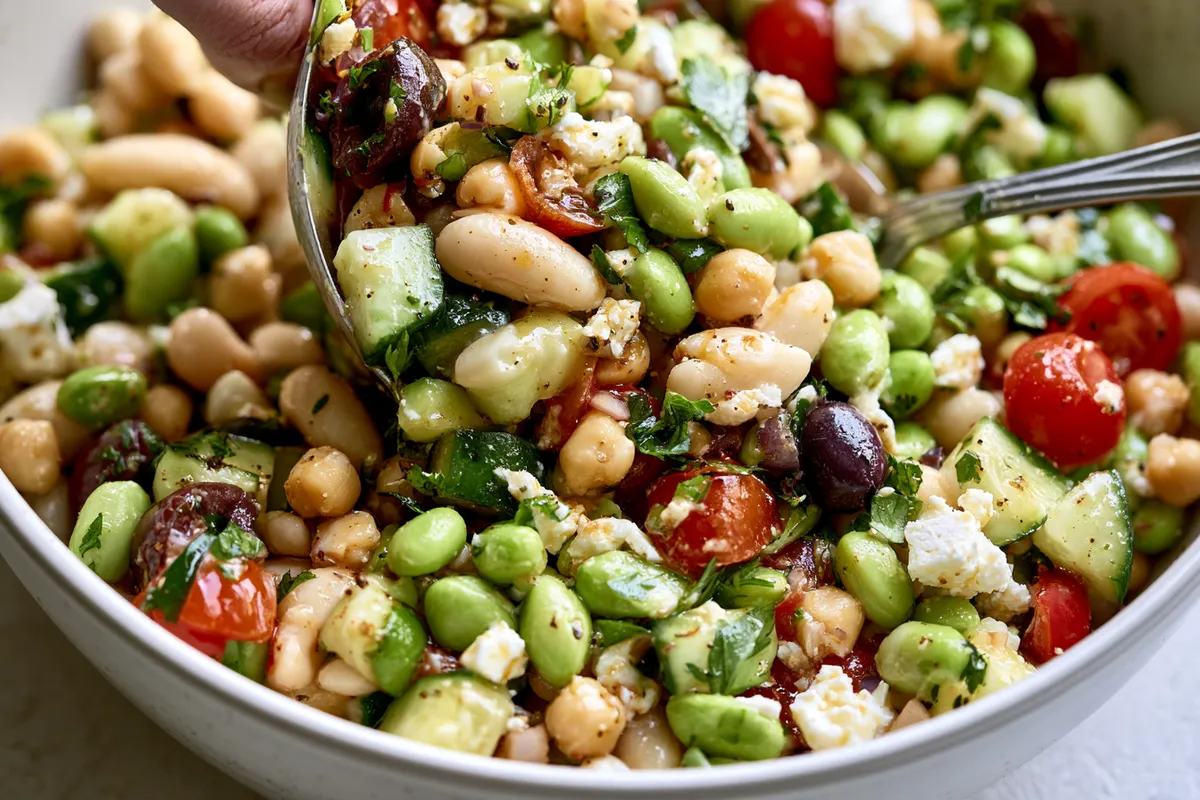 High Protein Bean Salad Meal Prep with Edamame and Feta 3 High protein bean salad meal prep being gently tossed in a large white bowl, with glossy lemon vinaigrette coating cannellini beans, chickpeas, edamame, feta, cucumber, tomatoes, olives, and parsley
